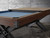 The Portland Slate Pool Table by Nixon Billiards is shown mid-transition with the optional dining top placed halfway across the playfield. This lifestyle photo captures the dual-purpose design in action, highlighting the contrast between the rustic hardwood cradle and the smooth, furniture-grade top surface. The matte black X-style steel legs ground the scene with industrial strength, while the remaining exposed Championship felt and racked Trophy Kit balls subtly reinforce that this table is game-night ready. A visual cue for versatility, shot exclusively for Sawyer Twain’s luxury game room collection.